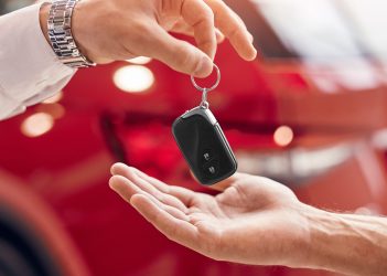 Unrecognizable client receiving keys of rent vehicle from manager against red vehicle in dealership